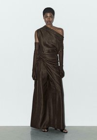 Brun, one-shoulder kjole med samlede detaljer på siden, lavet af tekstureret stof; parret med lange brune handsker og åbne sorte sandaler.