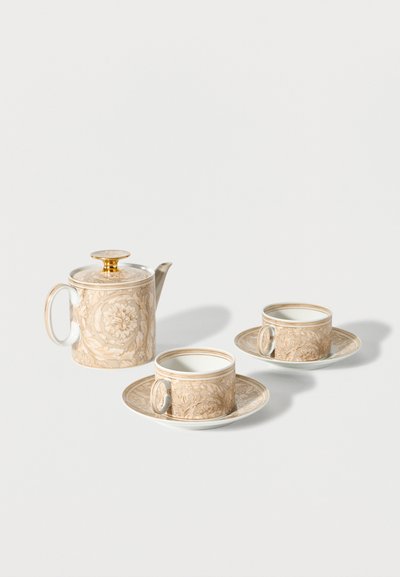 Beige floral patterned teapot with gold lid and two matching teacups on saucers, arranged on a plain white background.