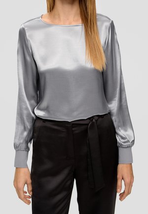 Femme portant un chemisier long brillant argenté et un pantalon noir taille haute avec une ceinture nouée à la taille, debout devant un fond clair.