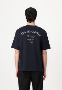 T-shirt en coton bleu marine avec un grand texte blanc au dos, portant la phrase « Café au Bord de la Plage » et d'autres détails. Coupe décontractée.