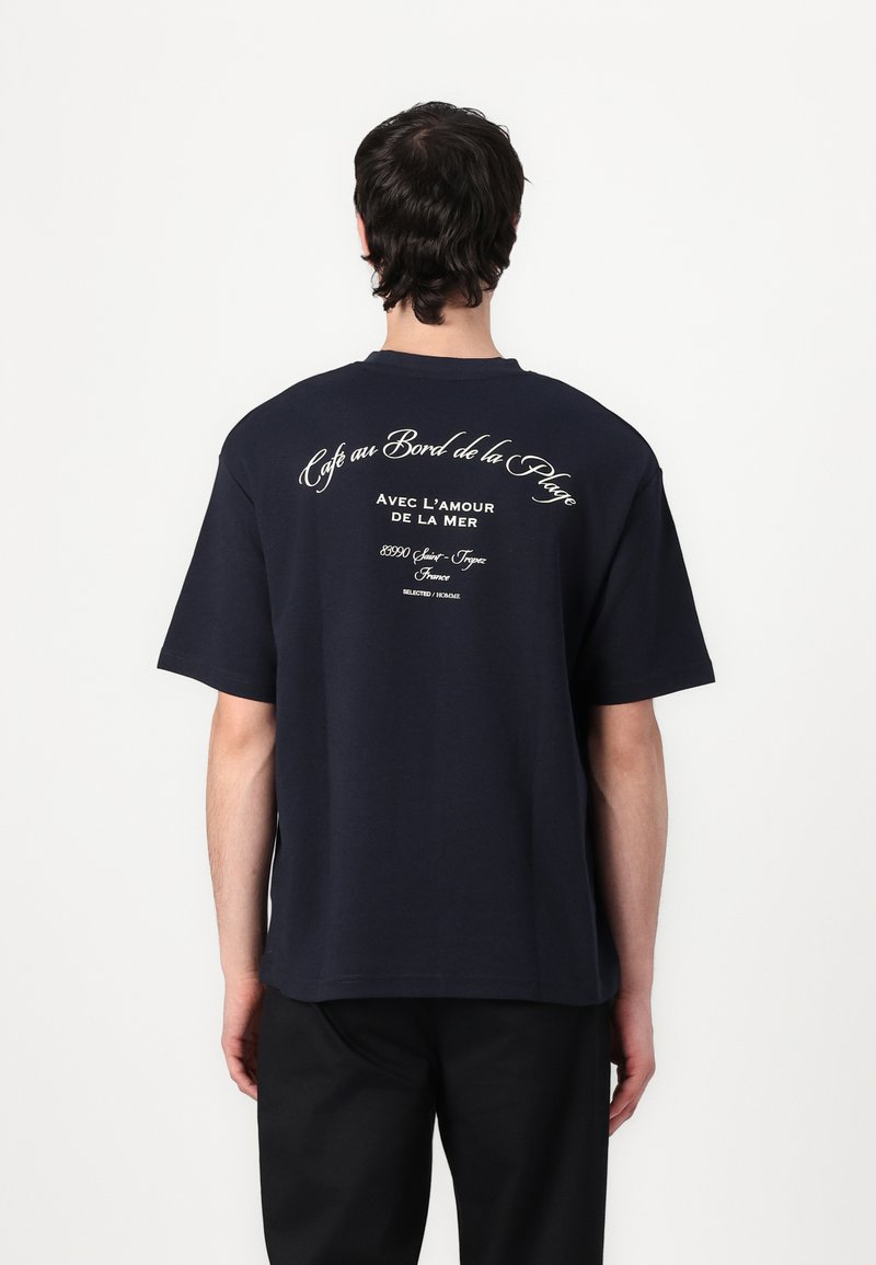 T-shirt en coton bleu marine avec un grand texte blanc au dos, portant la phrase « Café au Bord de la Plage » et d'autres détails. Coupe décontractée.