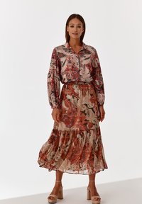 Langärmlige, bedruckte Bluse in Pastellpink mit floralem und paisley-artigem Muster, kombiniert mit einem gestuften, rüschierten Rock in erdigen Tönen.