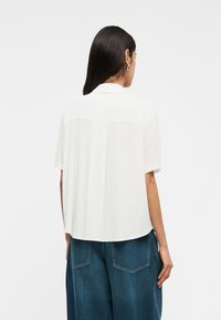 Γυναίκα με μακριά σκούρα μαλλιά που φοράει ασημένια σκουλαρίκια κρίκους, μια άνετη λευκή μπλούζα με κοντά μανίκια και μπλε τζιν με φαρδιά πόδια, που κοιτάζει προς τα πίσω σε απλό φόντο.