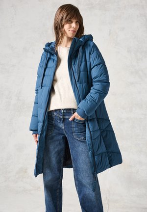 Manteau en teal rembourré avec capuche, doté d'une fermeture éclair et de poches, associé à un pull léger et à un jean en denim foncé.