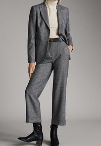 Costume gris sur mesure avec blazer structuré et pantalons larges. Tissu texturé, associé à un col roulé crème et des bottines noires.