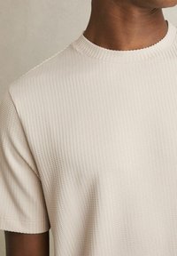Lys beige kortærmet t-shirt med ribbet struktur og rund halsudskæring, der har et glat, blødt stof og subtile mønsterdetaljer.