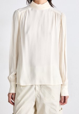 Vrouw die een ivoorkleurige blouse met lange mouwen, een hoge hals en geplooide details op de schouders en manchetten draagt, gecombineerd met lichtbeige broek.