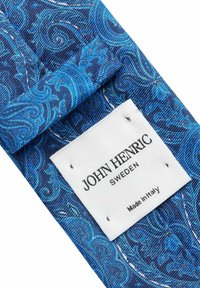 Sininen kuvioitu kangas, jossa on sileä pinta, ja siinä on pyörteitä. Valkoinen etiketti, jossa lukee "JOHN HENRIC SWEDEN Made in Italy", on kiinnitetty.