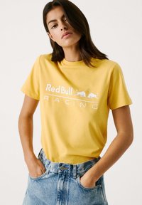 Gelbes Baumwoll-T-Shirt mit dem weißen "Red Bull Racing"-Logo, rundem Ausschnitt und kurzen Ärmeln, getragen mit blauen Jeans.