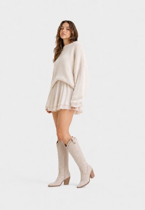 Donna che indossa un maglione crema oversize, gonna corta a strati e stivali beige al ginocchio con dettagli intagliati, in piedi su sfondo bianco.