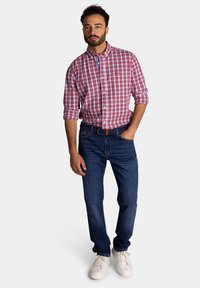 Rood-witte geblokte knoopshirt, donkerblauwe jeans en witte sneakers; model staat met handen in de zakken, neutrale achtergrond.