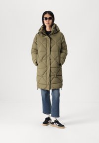 Olivgrüner, gesteppter Puffer-Mantel mit Kapuze, ausgestattet mit einem Frontreißverschluss, zwei Seitentaschen und einer lockeren Passform. Kombiniert mit ausgestellten Jeans und schwarzen Sneakers.