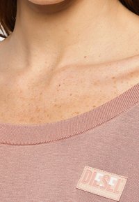 Różowa bluza z szerokim dekoltem i prążkowanymi krawędziami. Na przodzie znajduje się prostokątna metka z logo „DIESEL” o gładkiej teksturze.