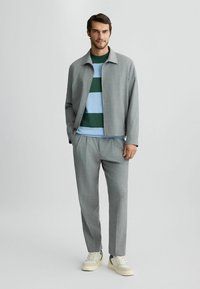 Hellgraue Anzugjacke mit Kragen, getragen über einem grün-blau gestreiften Pullover, kombiniert mit hellgrauen Hosen und cremefarbenen Sneakers.