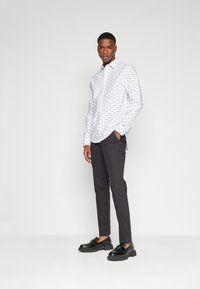 Camicia bianca con motivi di testo neri, abbinata a pantaloni grigio scuro e mocassini neri. La camicia ha una vestibilità comoda e un orlo curvo.