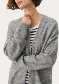 Cardigan gri tricotat cu o croi lejer, având mâneci lungi și o textură moale. Sub el, se vede o cămașă albă și albastră în dungi.