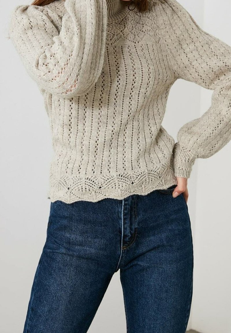 Femme portant un pull en maille beige avec des motifs en dentelle et un jean taille haute bleu foncé, posant avec une main sur la hanche et l'autre près du visage.