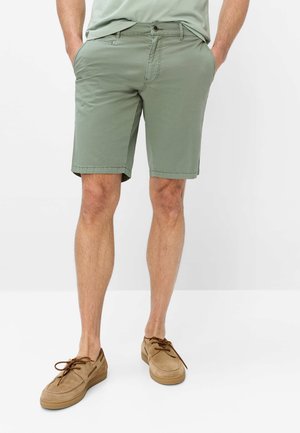 Mann trägt hellgrüne Shorts, beige Freizeitschuhe und hellgrünes Hemd mit Händen in den Taschen, steht vor weißem Hintergrund.