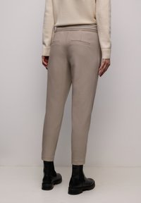 Person trägt beige schmal zulaufende Hose mit Gesäßpattentaschen, einen cremefarbenen Pullover und schwarze Stiefeletten und steht vor einem einfarbigen Hintergrund.