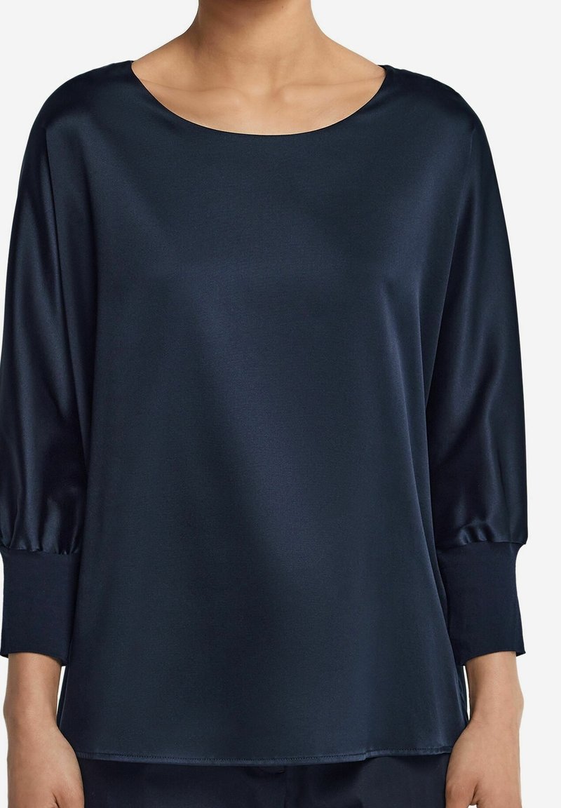 Femme portant un chemisier ample en satin bleu marine foncé avec manches trois-quarts et col rond sur un fond clair.