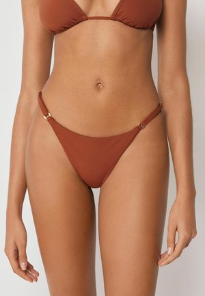 Kvinde iført bikini i rustfarve med justerbare metalringe på stropperne, der viser mave og øvre lår.