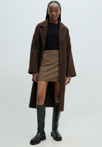 Brauner Wollmantel, schwarzes Oberteil, Minirock im Hahnentrittmuster und schwarze knielange Stiefel. Glatte Textur, entspannte Passform, mit großen Revers und Knöpfen.