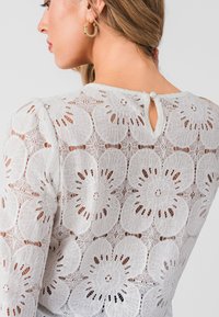 White lace top featuring a floral pattern, keyhole back detail with a button, and long sleeves. Textured fabric adds visual interest.