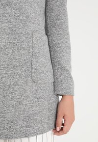 Grijze textured jas van zachte stof, met een enkele zak en lange mouwen. Het ontwerp heeft een schone, minimalistische snit.