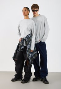 Cămașă gri cu mâneci lungi, pantaloni largi negri uzati, cămașă tartan verde legată la talie și botine negre cu talpă groasă.