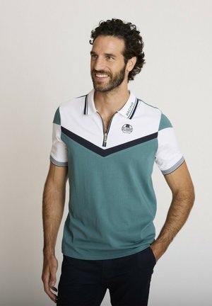 Homme aux cheveux bouclés et barbe portant un polo à manches courtes bleu sarcelle et blanc avec fermeture éclair et logo, debout avec une main dans la poche.