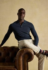 Mann mit Brille, navyblauem Langarmpolo, beigen Hosen und bunten gestreiften Socken sitzt lässig in einem braunen Lederarmstuhl.