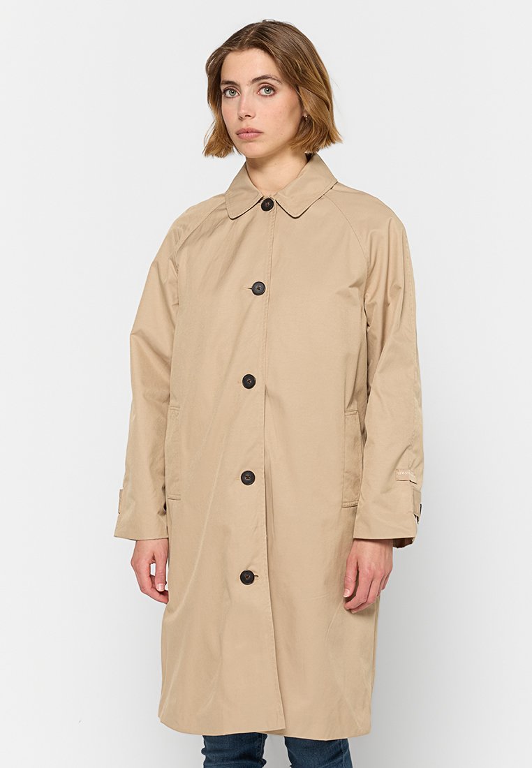 Donna con un trench beige a un bottone con bottoni neri, in piedi contro uno sfondo bianco uniforme.