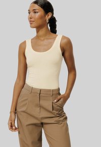 Un débardeur couleur crème avec un col festonné associé à un pantalon plissé beige clair, présenté sur un fond neutre.