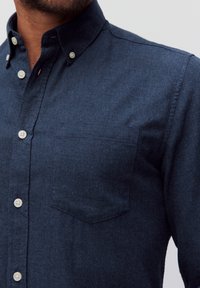 Donkerblauwe button-up shirt gemaakt van een gestructureerde stof. Bevat een kraag, witte knopen en een opgestikte zak op de linkerkant van de borst.