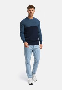 Pull en maille bleu marine et bleu avec un haut texturé et des panneaux inférieurs lisses, associé à un jean bleu clair et des baskets blanches.