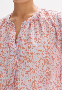 Blusa leggera e stampata in viola pastello e arancione. Presenta un collo a V, dettagli a pieghe e un tessuto testurizzato. Aspetto semi-trasparente.