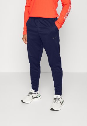 Personne portant un pantalon de jogging Nike bleu marine, des baskets Nike blanches et un t-shirt à manches longues orange, debout avec une main dans la poche, sur un fond uni.