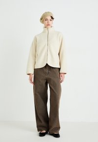 Cremefarbene Fleecejacke mit hohem Kragen, durchgehendem Reißverschluss und weiten Ärmeln, kombiniert mit lockeren braunen Hosen und einer gerippten beigen Beret.