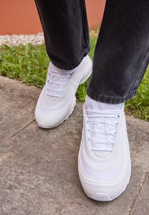 Personne portant des chaussures de sport blanches et un pantalon noir debout sur un chemin en béton avec de l'herbe à proximité.