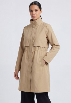 Vrouw die een beige lange jas draagt met hoge kraag en knopen aan de voorkant, met één hand in haar zak, staand tegen een effen lichte achtergrond.