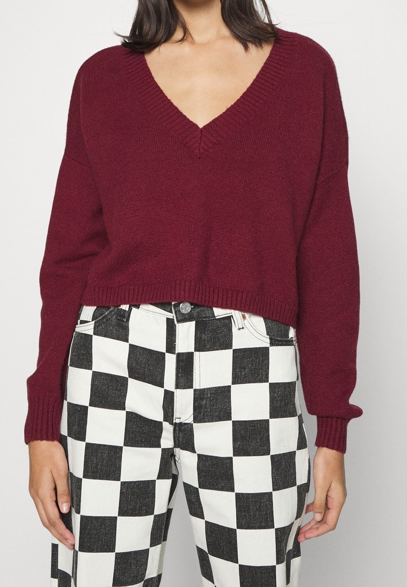 Femme portant un pull rouge foncé à col en V et un pantalon à motif damier noir et blanc, montrée de la poitrine aux hanches.