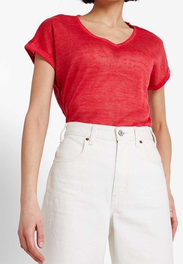 Donna che indossa una maglietta rossa trasparente a maniche corte con scollo a V, infilata in jeans bianchi a vita alta con tasche e chiusura a bottone.