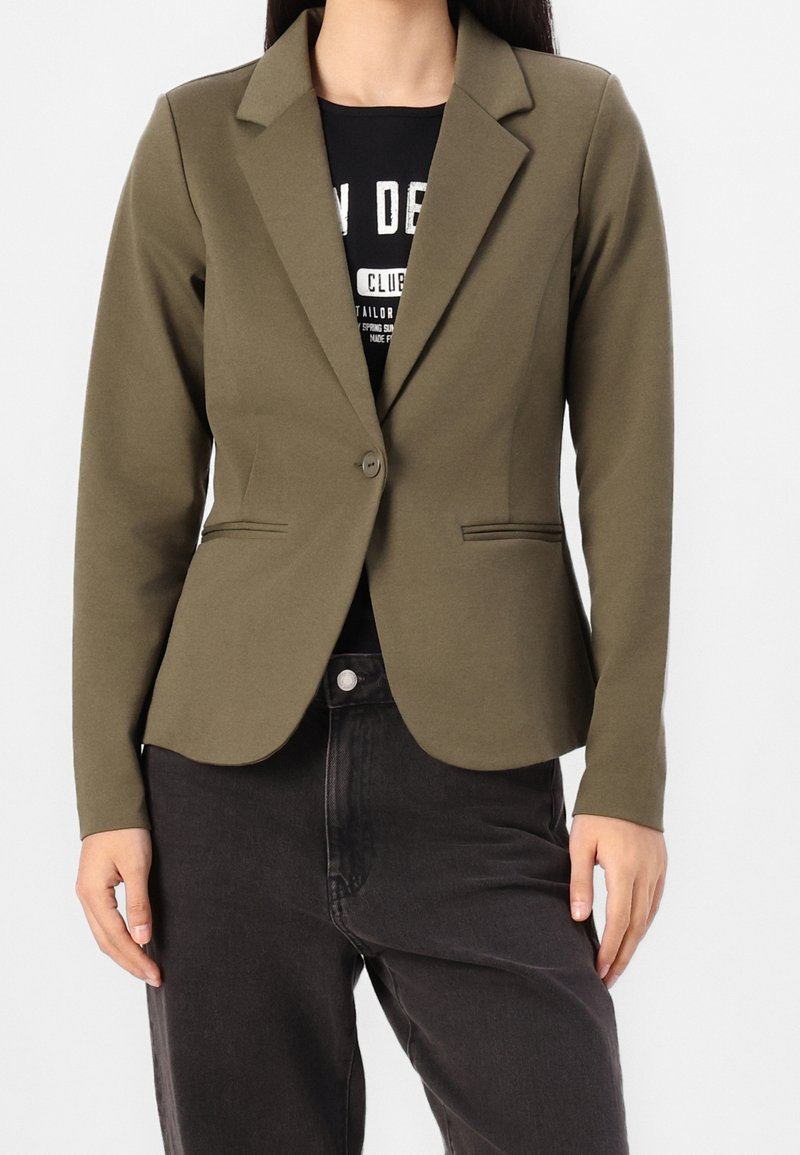 Femme portant un blazer vert olive à un seul bouton sur un t-shirt graphique noir et un jean gris foncé, debout devant un fond uni.