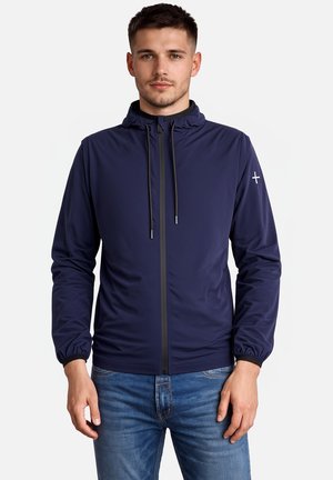 Mężczyzna ubrany w granatową kurtkę z kapturem, z zamkiem błyskawicznym z przodu i elastycznymi mankietami, w połączeniu z niebieskimi dżinsami, stojący na białym tle.