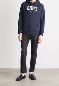 Persona vestida con una sudadera azul marino con el texto "GANT", vaqueros negros, calcetines blancos y mocasines negros de pie en un suelo gris liso contra una pared blanca.