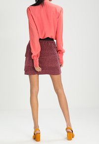 Blouse corail à manches longues, associée à une jupe magenta texturée avec des volants, et des chaussures compensées orange.