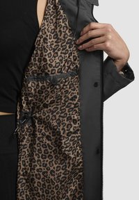 Veste extérieure noire avec doublure en imprimé léopard. L'intérieur comporte une poche à boutons et un cordon de serrage pour l'ajustement.