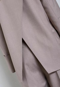 Svetlo taupe sako so šitým oblekom z textúrovaného materiálu, so striktnými líniami a jedným vreckom na saku, predstavujúc moderné dizajnové prvky.