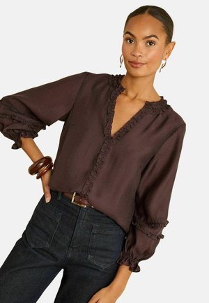 Dunkelbraune Bluse mit gerafftem Ausschnitt und Bündchen, mit V-Ausschnitt. Kombiniert mit dunklen Jeans, die Vorderhosentaschen zeigen.