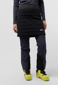 Jupe noire isolée avec un motif matelassé horizontal, associée à un pantalon gris foncé et des bottes de ski jaune vif. Détail du logo sur la jupe.